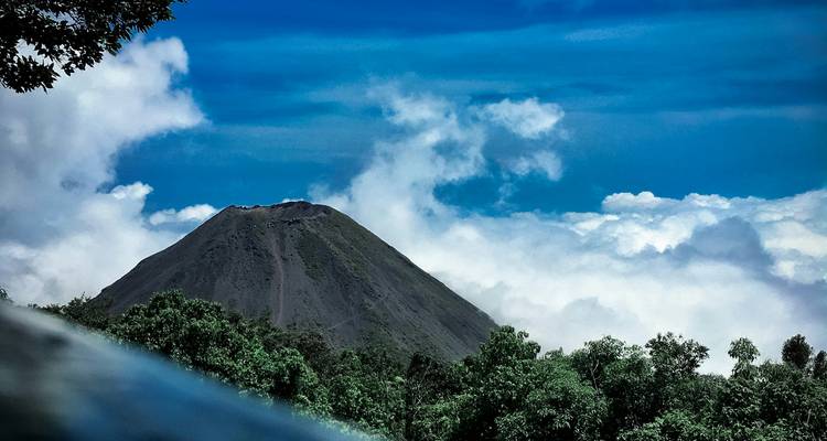 Conical volcano rises above a sea of clouds framed by lush tropical forest.