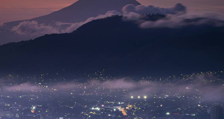 Twinkling city lights beneath misty hills with a silhouetted volcano at dusk.