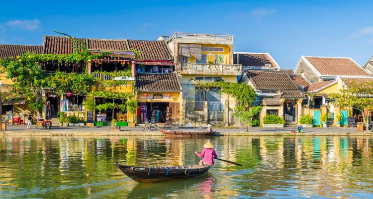 Wooden sampan with conical-hatted rower glides along Hoi An’s colorful riverside old town.