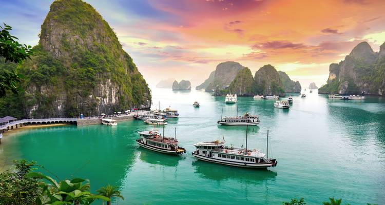 Ha Long Bay limestone karsts rise from emerald water as tour boats float beneath a vibrant sunset sky.