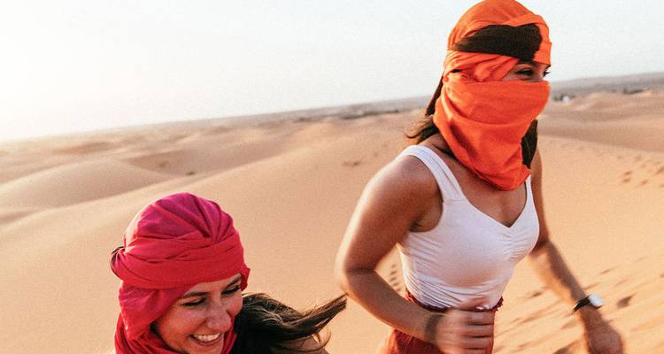 Two women in colourful scarves run across golden desert dunes at sunset.
