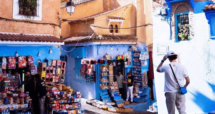 Colorful street market in the blue-painted medina with stalls of magazines and souvenirs, tourist taking a photo.