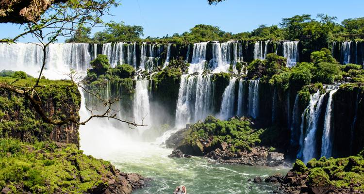 Thunderous multi-tiered Iguazu Falls cascade into a misty gorge where a small speedboat braves the swirling water below.