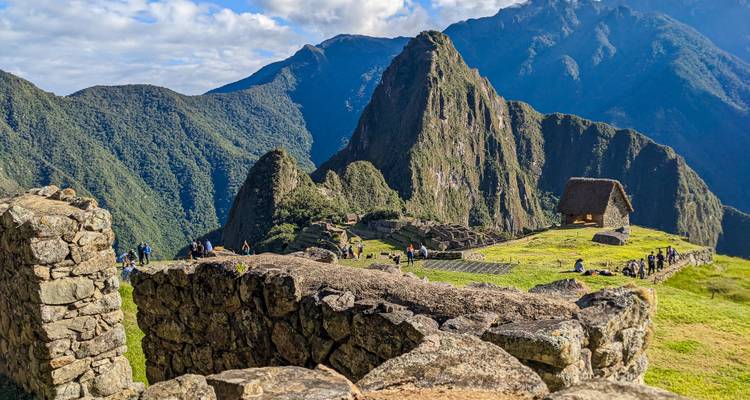 Bezoekers wandelen langs stenen muren en grasrijke terrassen bij Machu Picchu met een rieten wachthuis en torenhoge bergtoppen op de achtergrond.