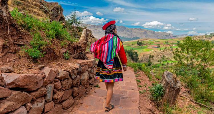 Mujer con ropa tradicional andina vibrante caminando por un sendero de piedra que da vista a un valle terrazado y exuberante, con las montañas andinas a lo lejos bajo un cielo despejado.