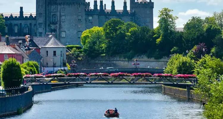 Small red boat with two occupants travels along a flower-lined river below a stone bridge, with a medieval castle and lush greenery behind.