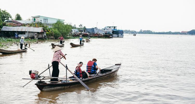 Un rameur local portant un chapeau conique propulse un petit bateau en bois transportant des touristes équipés de gilets de sauvetage le long d'une large rivière boueuse avec des maisons riveraines et d'autres sampans en arrière-plan.