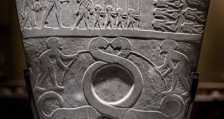 Close-up of an ancient Egyptian ceremonial palette displayed in a museum, showing intertwined serpents and armed figures.