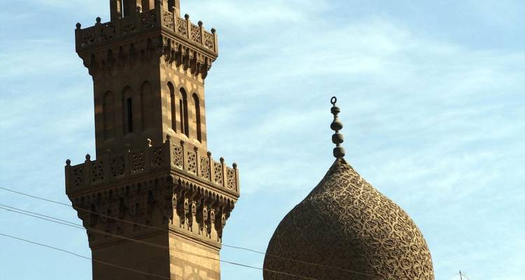 Detailed close-up of a patterned stone minaret and domed roof set against a pale blue sky.