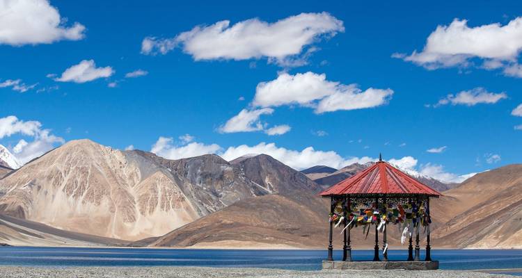 Gazebo draped in prayer flags beside pristine blue Pangong Lake with dramatic mountain backdrop.