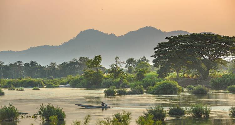 Ein kleines Motorboot gleitet sanft einen ruhigen Fluss entlang, der von üppigen Bäumen gesäumt wird, während im Hintergrund nebelverhüllte Berge sich bei Sonnenuntergang abzeichnen.
