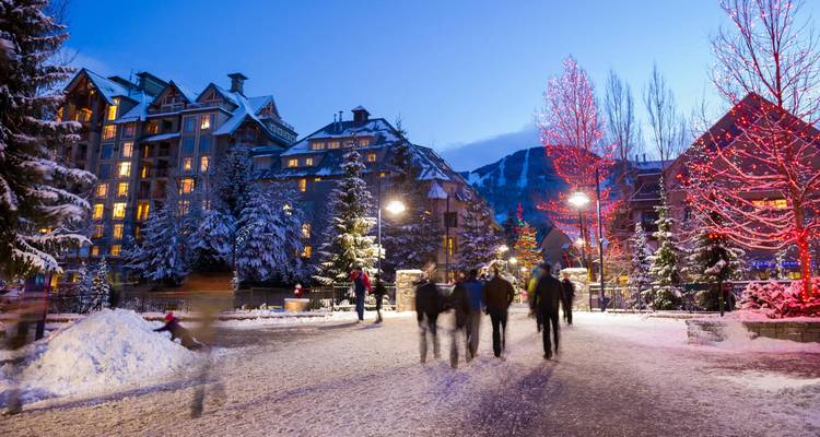 Snow-covered pedestrian village with people strolling under evening lights and festive trees.