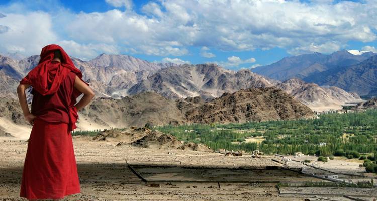 Red-robed monk overlooks a broad arid valley ringed by stark Himalayan mountains under scattered clouds.