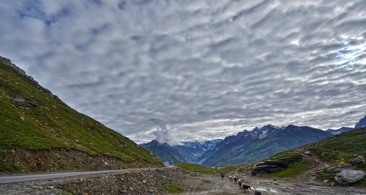 Mountain road winding through green valley beneath dramatic textured cloud blanket in the Himalayas.