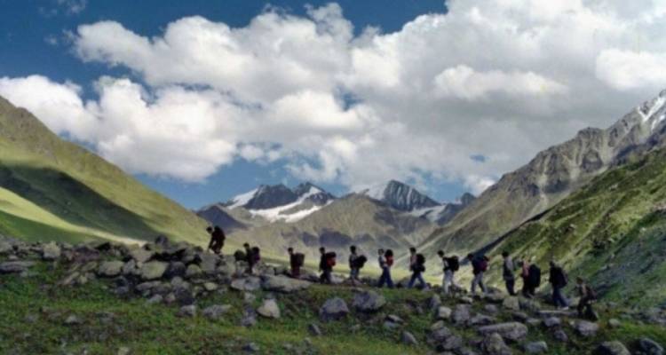 Reihe von Wanderern, die eine felsige Wiese überqueren, mit aufragenden schneegestreiften Gipfeln im Hintergrund.