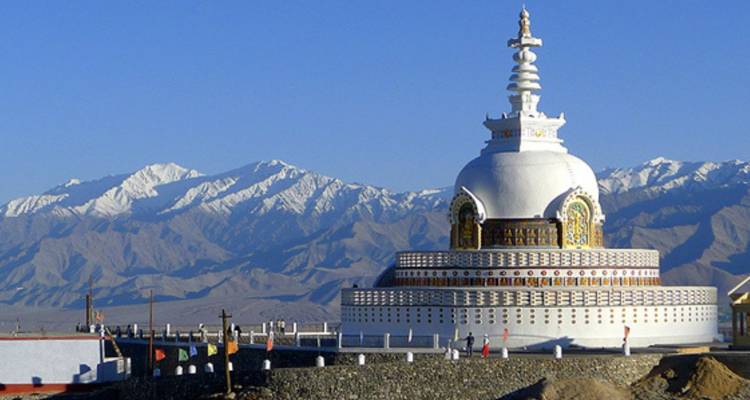 Weiße kuppelförmige Shanti Stupa vor zerklüfteter Himalaya-Bergkette und tiefblauem Himmel.