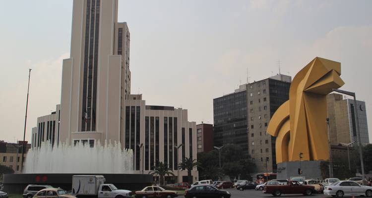 View of the golden modernist Caballito sculpture beside tall buildings and a fountain.