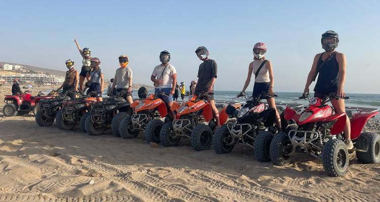 Group of riders pose on red and orange quad bikes on a sandy beach with the sea in the background.