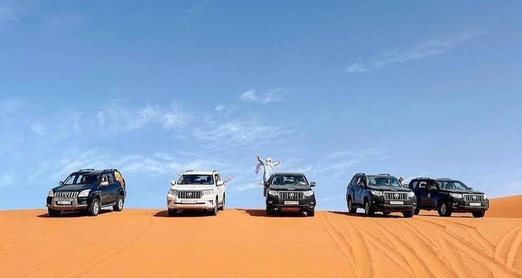Five off-road vehicles line a dune ridge while passengers joyfully jump against a blue sky.