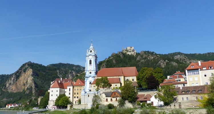 Charming riverside village of Dürnstein with blue church tower backed by forested hills and castle ruins under clear sky.