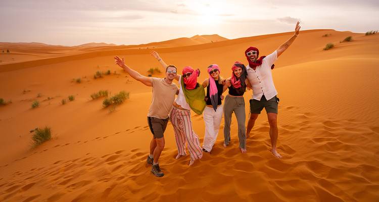 Cinq amis portant des foulards colorés célèbrent sur les dunes dorées du Sahara sous un ciel chaud du soir.