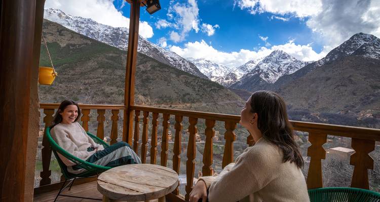 Deux femmes se détendent sur un balcon en bois donnant sur les montagnes de l'Atlas enneigées et une vallée verdoyante.