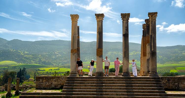 Les visiteurs se tiennent parmi les hautes colonnes romaines de Volubilis avec la campagne verdoyante vallonnée en arrière-plan.
