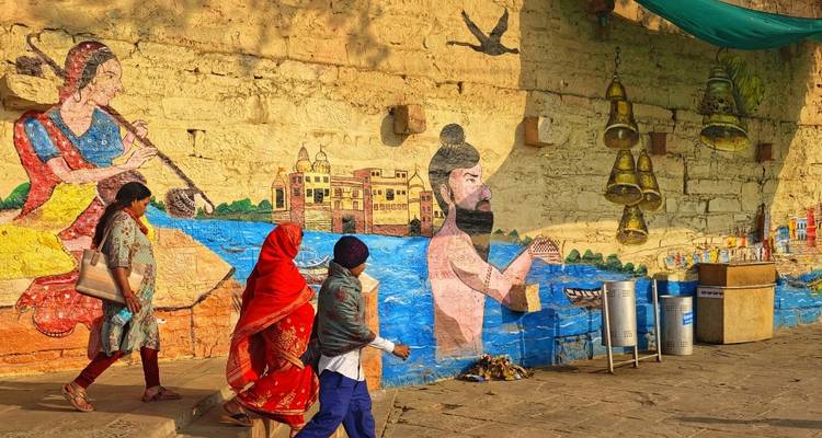 Lokale bewoners in kleurrijke kleding lopen langs levendige muurschilderingen aan de rivieroever die de Indiase cultuur uitbeelden.