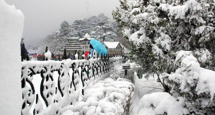 Snow-covered promenade in Shimla with wrought-iron railing and people holding umbrellas.