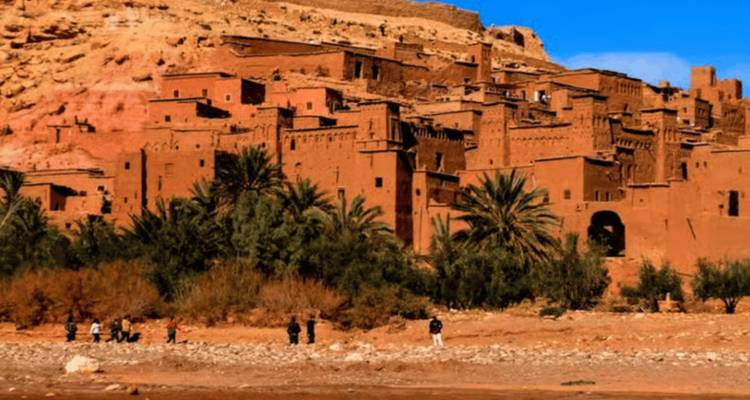 Die Terrakotta-Gebäude von Ait Benhaddou, eingerahmt von Palmen und felsigem Hang unter blauem Himmel.