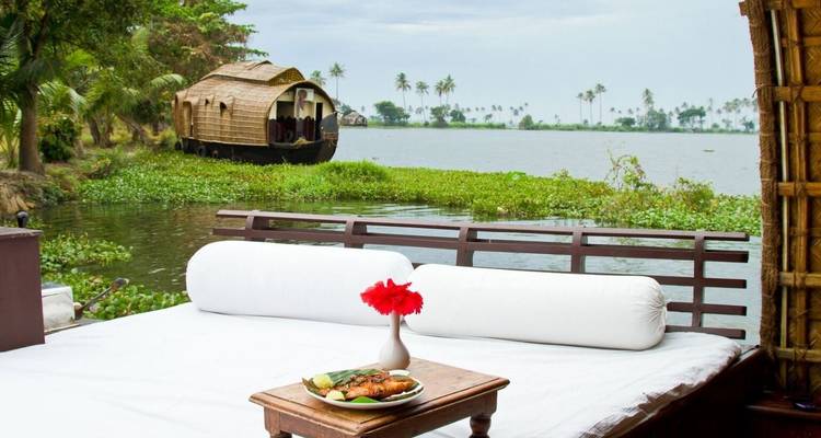 Un lit de repos confortable sur le pont d'une péniche surplombe les backwaters du Kerala avec un autre bateau au toit de chaume qui navigue au loin.