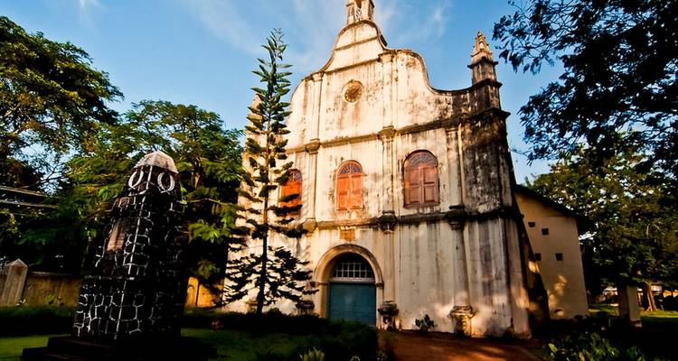 Historic St. Francis Church with weathered white façade, tall arched windows and a unique bell-topped gable amid greenery