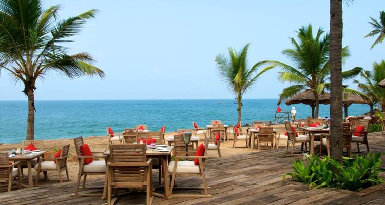 Open-air beach restaurant with wooden tables and red napkins facing the blue sea under clear sky