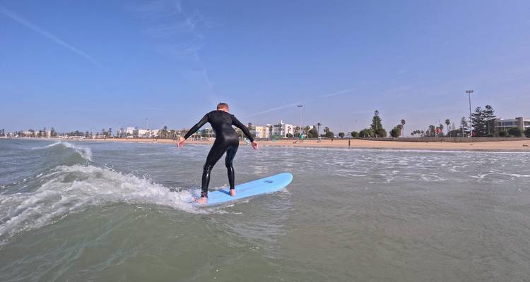 Surfer im Neoprenanzug reitet eine kleine Welle in Richtung des sandigen Stadtstrandes unter klarem blauem Himmel.