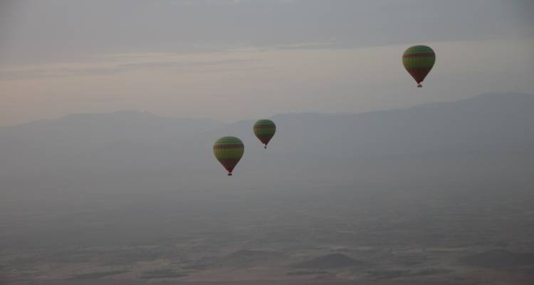 Drei bunte Heißluftballons schweben lautlos in einem dunsigen Morgenhimmel über fernen Bergen.