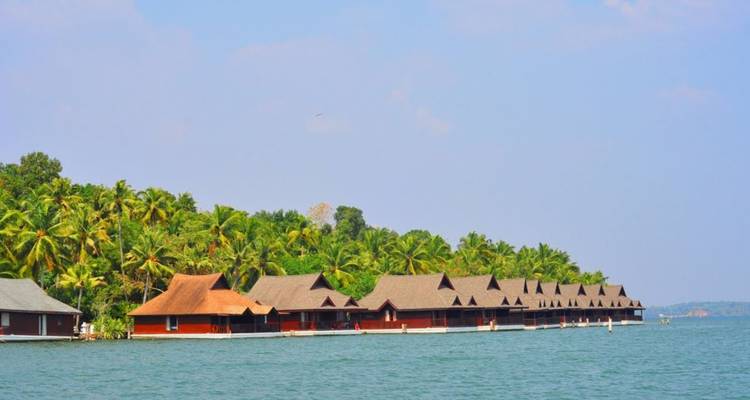 Row of rustic over-water cottages with thatched roofs lining a calm backwater lagoon bordered by palm trees.