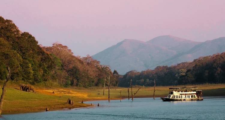 Serene lake with tour boat passing forested hills and hazy mountains in gentle evening light.