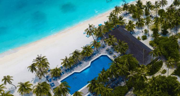 Aerial shot of a beachfront resort with palm-lined pool, white sand and vivid turquoise lagoon in the Maldives.