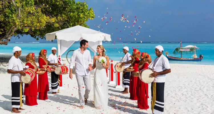 Beach wedding ceremony with bride and groom walking through confetti petals accompanied by traditional Maldivian drummers in red robes.