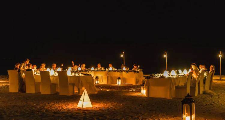 Long candlelit table set for an intimate dinner on the sandy beach at night, guests enjoying the evening glow.