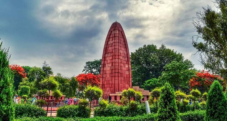 The red sandstone spire of Jallianwala Bagh memorial rises amid lush gardens under dramatic clouds.