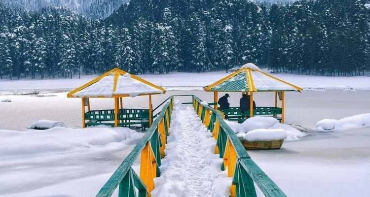 A snow-blanketed pier with colorful gazebos leads onto a frozen alpine lake surrounded by pine forests.