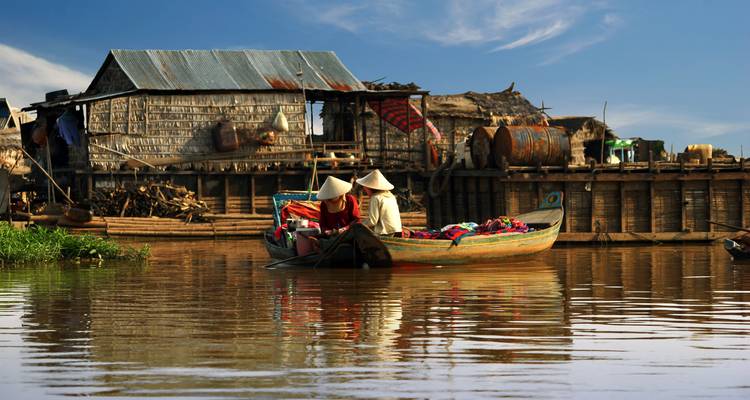 Los lugareños con sombreros cónicos reman una embarcación de madera pasando por casas sobre pilotes en un río marrón y tranquilo
