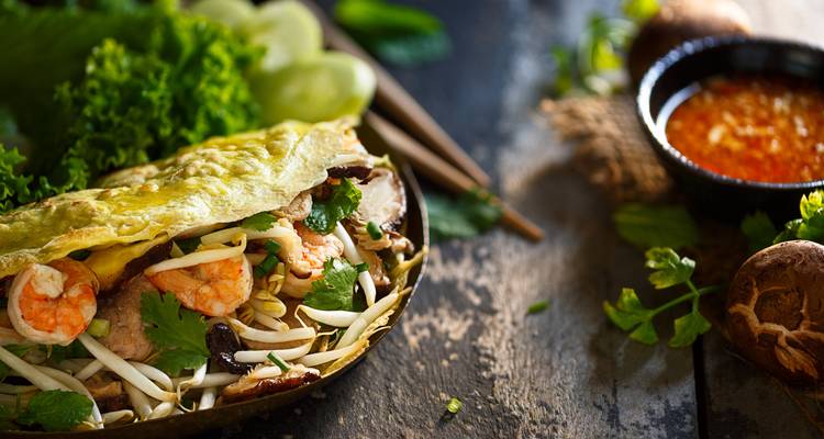 Nahaufnahme eines vietnamesischen knusprigen Pfannkuchens gefüllt mit Garnelen, Pilzen und Sojasprossen neben frischen Kräutern