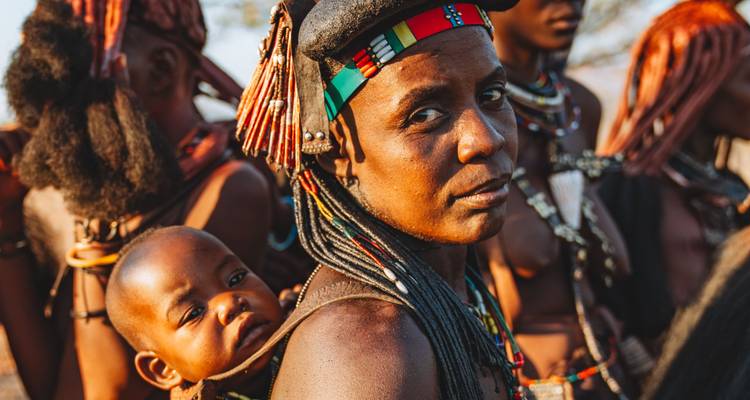 Mère himba portant un bébé sur son dos, toutes deux parées d'ornements traditionnels parmi les compagnons du village