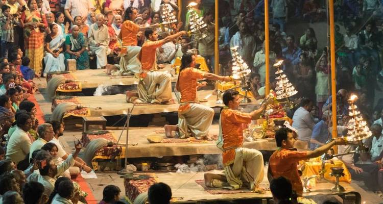 Priester in orangefarbenen Roben führen eine aufwendige Abend-Aarti mit flammenden Lampen auf, während eine große Menschenmenge zuschaut.
