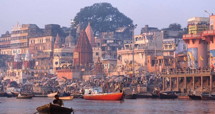 Lebhafte Flussuferpromenade von Varanasi mit bunten Tempeln, Ghats, Booten und Pilgern entlang des Ganges bei Morgendämmerung.