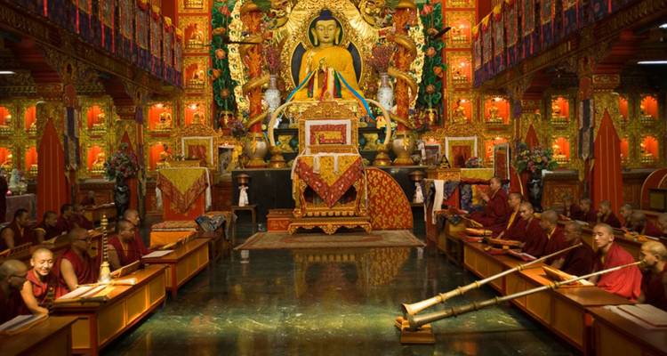 Mönche in roten Gewändern versammelt in einer reich verzierten Klosterhalle mit einer goldenen Buddha-Statue.