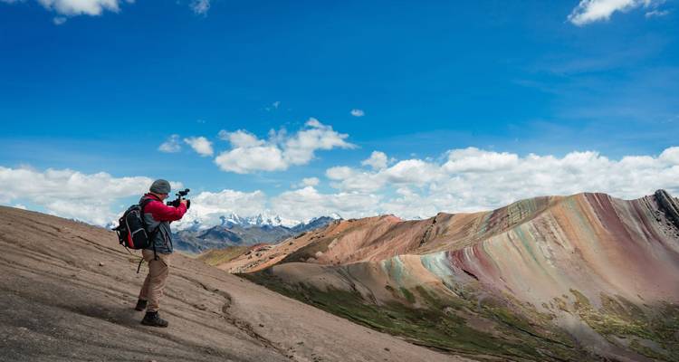 Hiker photographs Peru’s Rainbow Mountain ridge under a brilliant sky on a steep slope.