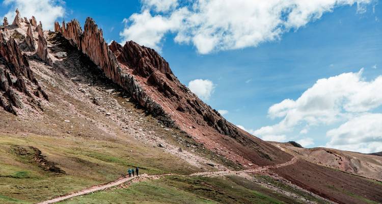 Sharp fin-like rock formations rise above grassy slopes where tiny hikers follow a winding trail.